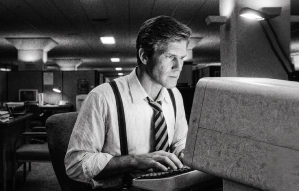Grainy black and white photograph of a man in shirt, tie and suspenders at a CRT computer in an 1980s office, lit by a single desk lamp.